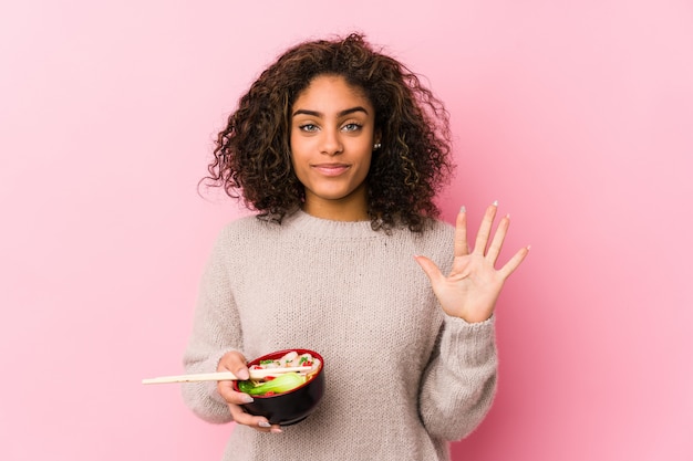 Joven mujer afroamericana comiendo fideos sonriendo alegre mostrando número cinco con los dedos.