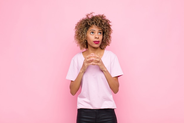 Joven mujer afroamericana conspirando y conspirando, pensando trucos y trampas astutos, astuto y traicionando contra la pared rosa