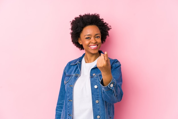 Joven mujer afroamericana contra un fondo rosa aislado apuntando con el dedo como si invitara a acercarse.