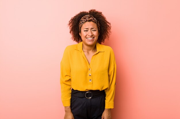 Joven mujer afroamericana contra una pared de color rosa se ríe y cierra los ojos, se siente relajado y feliz.