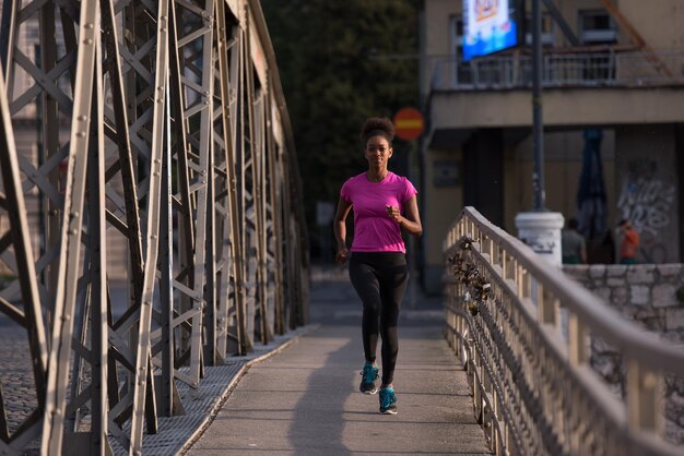 Joven mujer afroamericana deportiva corriendo en la acera cruzando el puente temprano en la mañana trotando con la escena del amanecer de la ciudad en el fondo