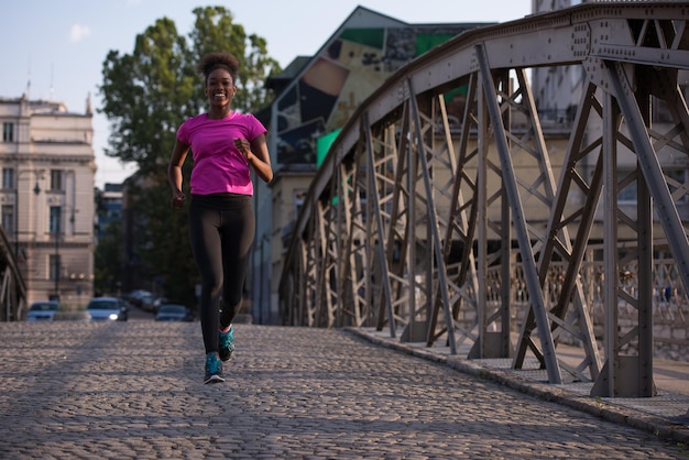 Joven mujer afroamericana deportiva corriendo en la acera cruzando el puente temprano en la mañana trotando con la escena del amanecer de la ciudad en el fondo
