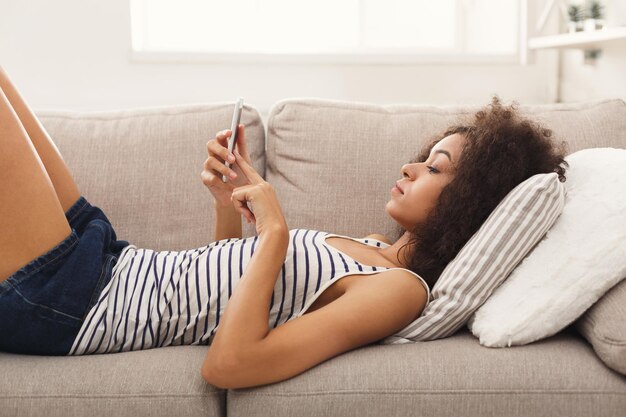 Joven mujer afroamericana enviando mensajes en el teléfono inteligente en casa, cómodamente acostado en el sofá beige, copie el espacio, vista lateral