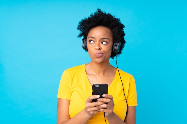 Joven mujer afroamericana escuchando música con un móvil sobre pared azul aislado