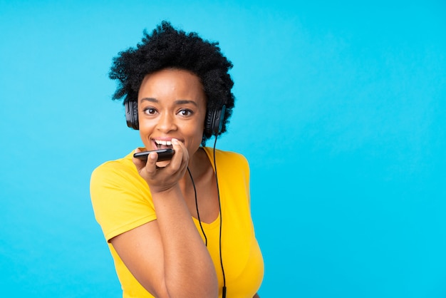 Joven mujer afroamericana escuchando música con un móvil sobre pared azul aislado