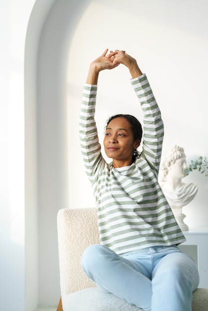 Joven mujer afroamericana estirándose mientras está sentada mirando la ventana por la mañana