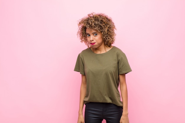 Joven mujer afroamericana con una expresión tonta, loca, sorprendida, hinchando las mejillas, sintiéndose rellena, gorda y llena de comida contra la pared rosa