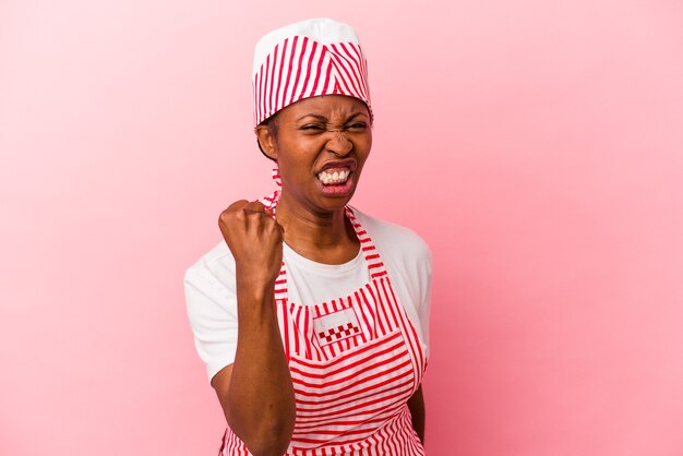 Joven mujer afroamericana fabricante de helados aislada sobre fondo rosa mostrando el puño a la cámara, expresión facial agresiva.