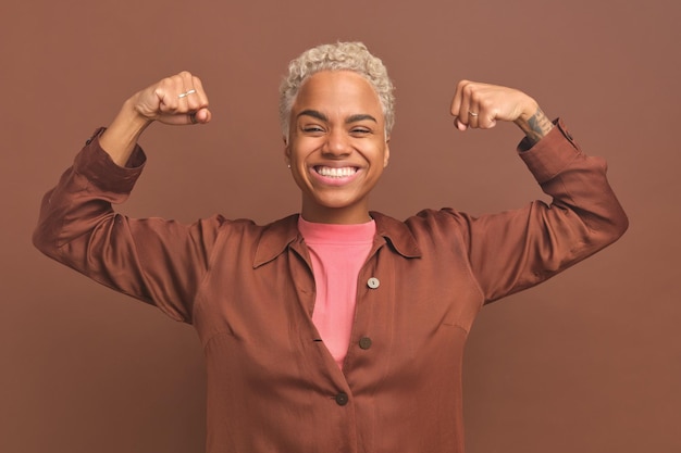 Joven mujer afroamericana feliz mostrando bíceps se encuentra sobre fondo marrón