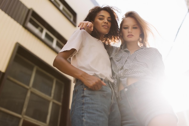 Joven mujer afroamericana fresca con cabello rizado oscuro en camiseta y hermosa mujer con cabello rubio en camisa mirando soñadoramente a la cámara mientras pasan tiempo juntos al aire libre