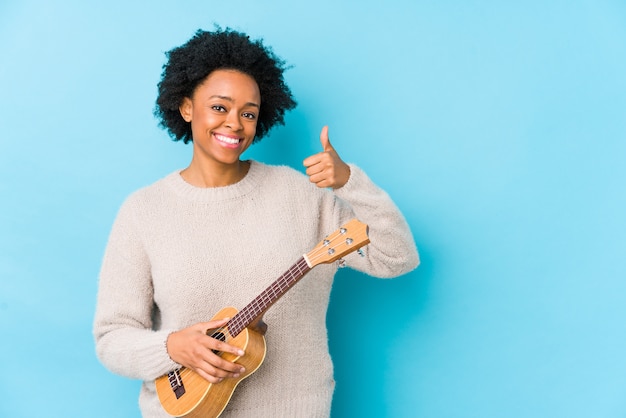 Joven mujer afroamericana jugando ukelele aislado sonriendo y levantando el pulgar hacia arriba