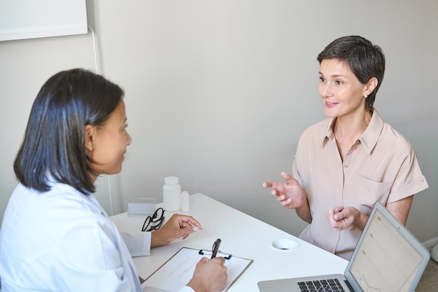 Joven mujer afroamericana médico médico anotando los síntomas del paciente de mediana edad