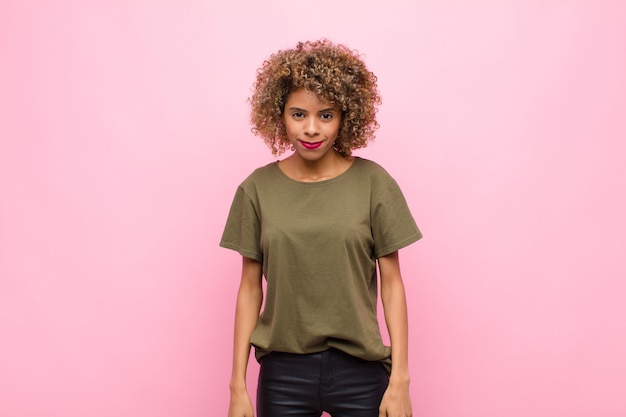 Joven mujer afroamericana mirando orgulloso, confiado, fresco, descarado y arrogante, sonriendo, sintiéndose exitoso contra la pared rosa