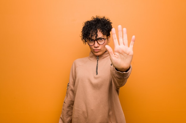 Joven mujer afroamericana mixta contra una pared marrón de pie con la mano extendida que muestra la señal de stop, impidiéndole.