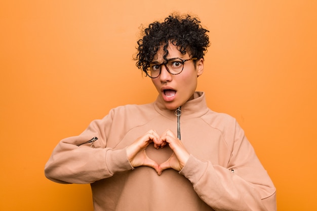 Joven mujer afroamericana mixta contra una pared marrón sonriendo y mostrando una forma de corazón con las manos.