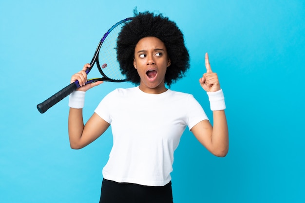 Joven mujer afroamericana en la pared azul jugando tenis y apuntando hacia arriba