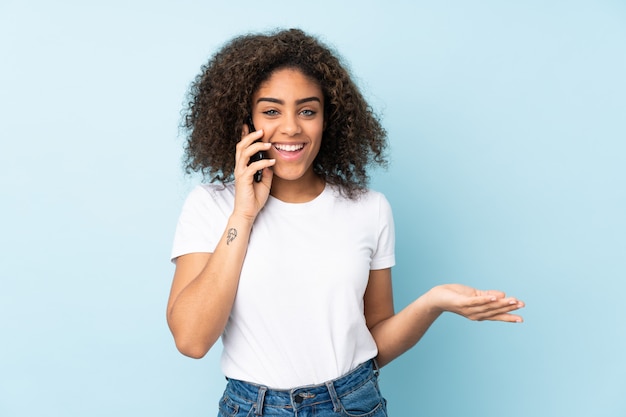 Joven mujer afroamericana en la pared azul manteniendo una conversación con el teléfono móvil con alguien