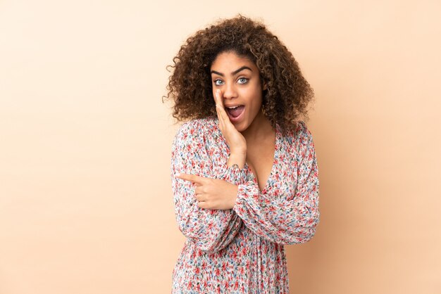 Joven mujer afroamericana en pared beige apuntando hacia un lado para presentar un producto y susurrando algo