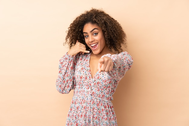 Joven mujer afroamericana en pared beige haciendo gesto de teléfono y señalando