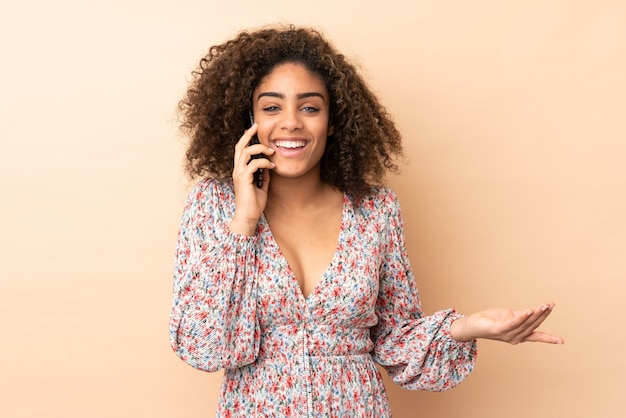 Joven mujer afroamericana en pared beige manteniendo una conversación con el teléfono móvil con alguien