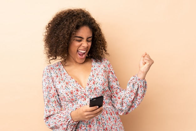 Joven mujer afroamericana en pared beige con teléfono en posición de victoria