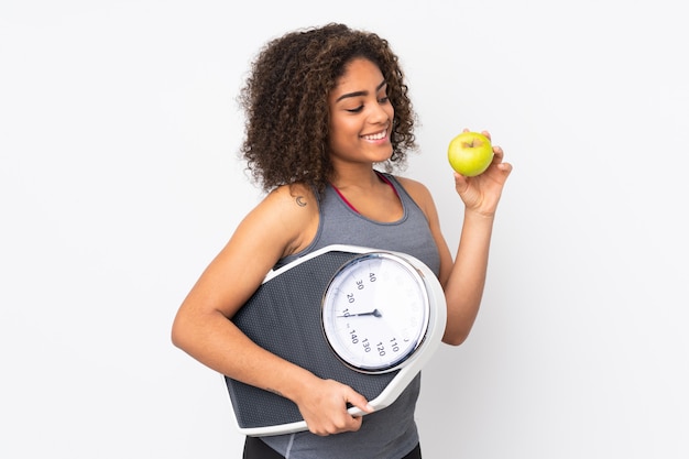 Joven mujer afroamericana en pared blanca con máquina de pesaje y con una manzana