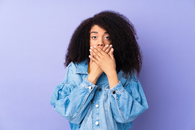 Joven mujer afroamericana en pared púrpura cubriendo la boca con las manos