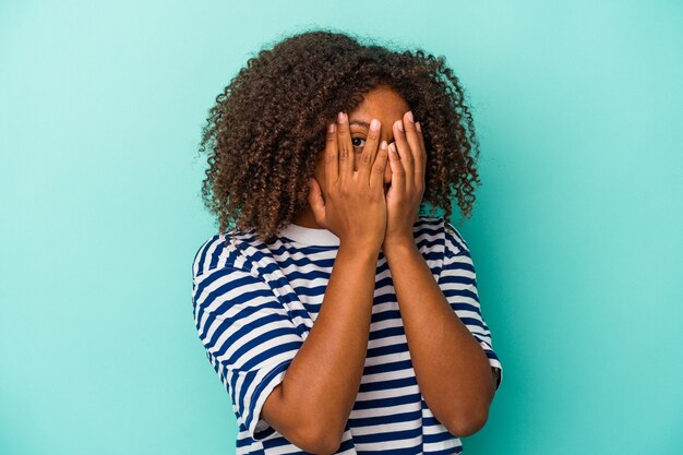 Joven mujer afroamericana con pelo rizado aislado sobre fondo azul parpadea a través de los dedos asustada y nerviosa.