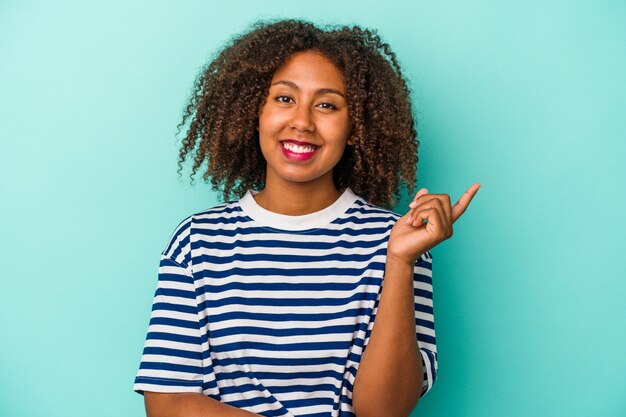 Joven mujer afroamericana con pelo rizado aislado sobre fondo azul sonriendo alegremente apuntando con el dedo índice.