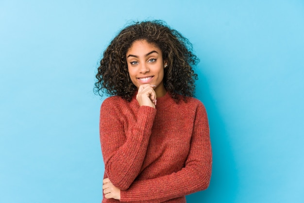 Joven mujer afroamericana de pelo rizado sonriendo feliz y confiada, tocando la barbilla con la mano.