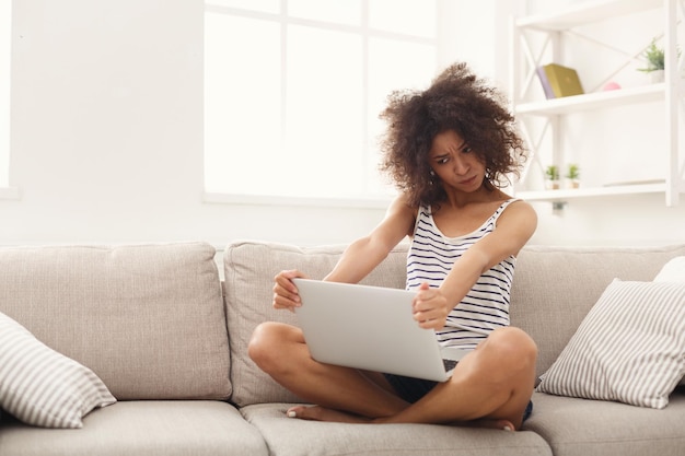 Joven mujer afroamericana preocupada que trabaja en la computadora portátil sentada en un sofá beige, red de surf, espacio de copia