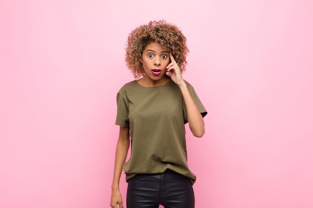 Joven mujer afroamericana que parece sorprendida, con la boca abierta, sorprendida, al darse cuenta de un nuevo pensamiento, idea o concepto contra la pared rosa