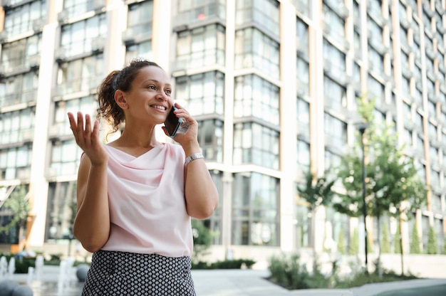 Joven mujer afroamericana de raza mixta hablando por teléfono móvil en el fondo de los altos edificios urbanos. Conceptos comerciales y de comunicación