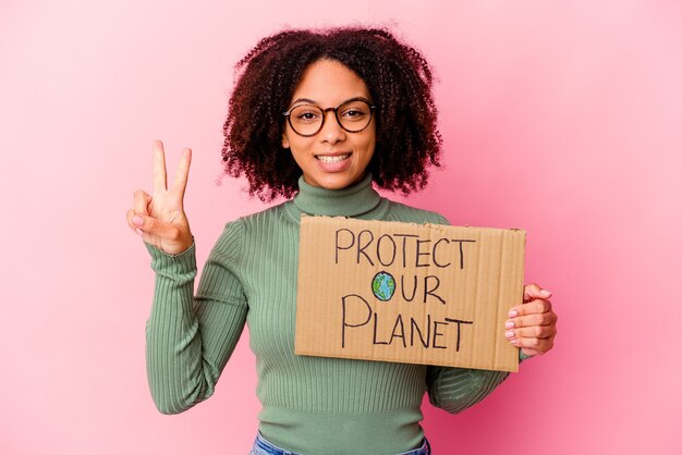 Joven mujer afroamericana de raza mixta sosteniendo un cartón de proteger nuestro planeta mostrando el número dos con los dedos.