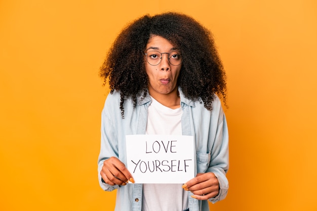 Joven mujer afroamericana rizada sosteniendo un cartel de amor a sí mismo se encoge de hombros y abre los ojos confundidos.