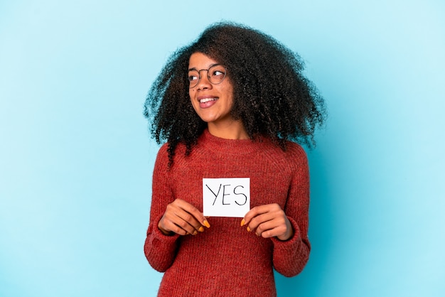 Joven mujer afroamericana rizada sosteniendo un cartel de sí mira a un lado sonriente, alegre y agradable.