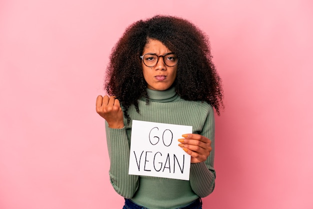 Joven mujer afroamericana rizada sosteniendo un cartel vegano que muestra el puño, expresión facial agresiva.