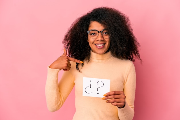 Joven mujer afroamericana rizada sosteniendo un interrogatorio en una persona de cartel apuntando con la mano a un espacio de copia de camisa, orgulloso y seguro