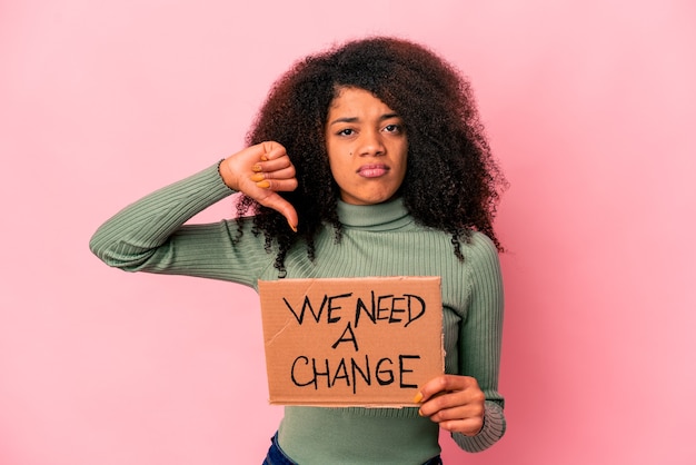 Joven mujer afroamericana rizada sosteniendo un necesitamos un cartón de cambio mostrando un gesto de aversión, pulgares hacia abajo. Concepto de desacuerdo.