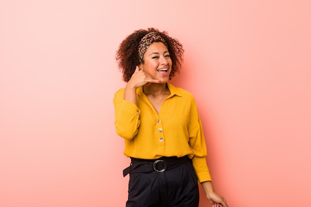 Joven mujer afroamericana en rosa mostrando un gesto de llamada de teléfono móvil con los dedos.