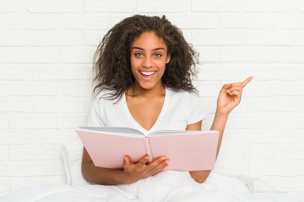 Joven mujer afroamericana sentada en la cama estudiando sonriendo alegremente señalando con el dedo lejos.