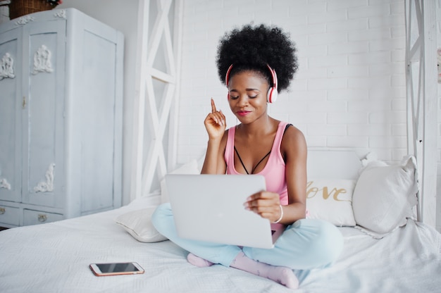 Joven mujer afroamericana sentada en la cama mientras trabajaba en la computadora portátil y escucha música en los auriculares. Ella levantó su dedo hacia arriba.