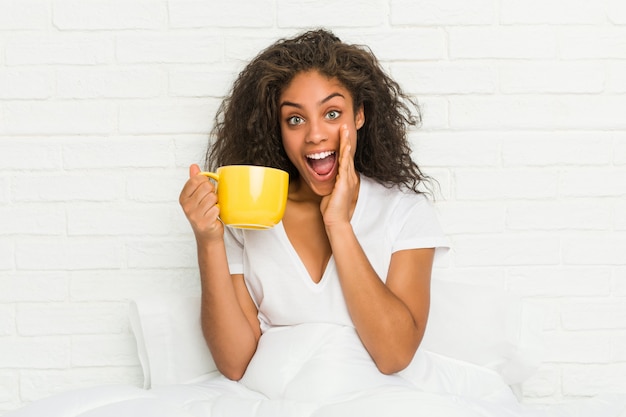 Joven mujer afroamericana sentada en la cama sosteniendo una taza de café gritando emocionado al frente.