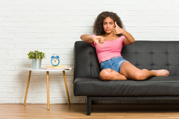 Joven mujer afroamericana sentada en el sofá mostrando los pulgares hacia arriba y hacia abajo, difícil elegir el concepto