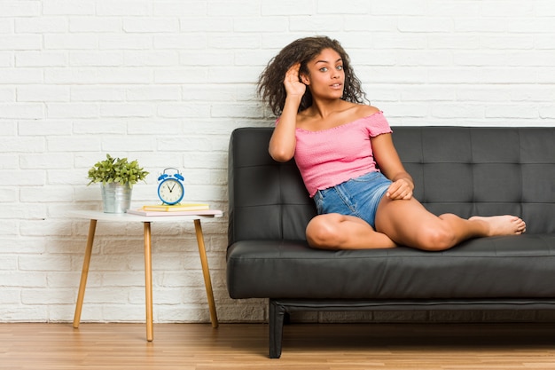 Joven mujer afroamericana sentada en el sofá tratando de escuchar un chisme.