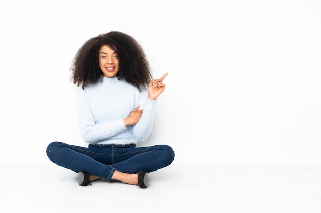 Joven mujer afroamericana sentada en el suelo apuntando con el dedo al lado