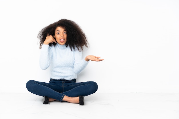 Joven mujer afroamericana sentada en el suelo haciendo gesto de teléfono y dudando