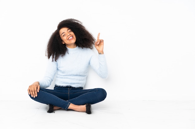 Joven mujer afroamericana sentada en el suelo mostrando y levantando un dedo en señal de lo mejor