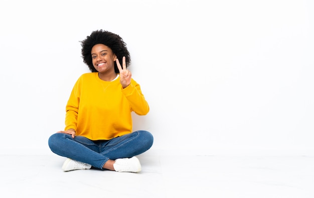 Joven mujer afroamericana sentada en el suelo sonriendo y mostrando el signo de la victoria