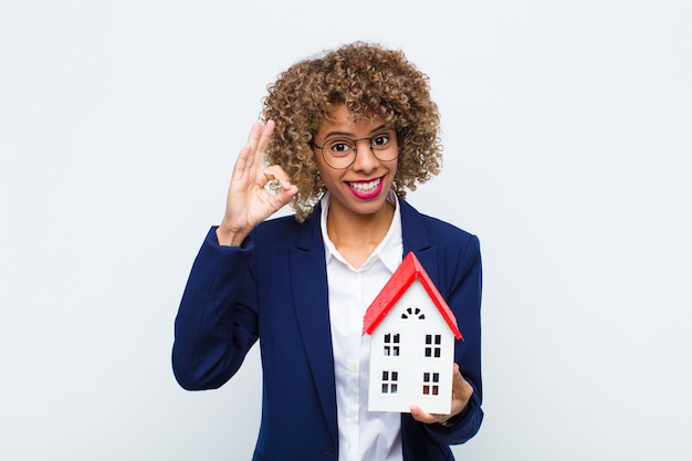 Joven mujer afroamericana se siente feliz, relajada y satisfecha, mostrando aprobación con gesto bien, sonriendo con la casa modelo